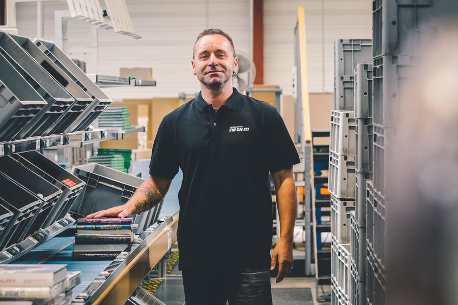 Arvato employee stands at a packing station and looks proudly into the camera