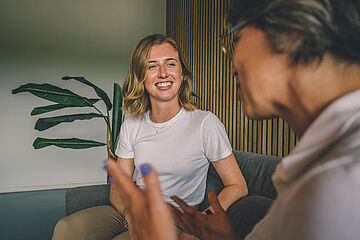 Zwei Mitarbeiterinnen unterhalten sich auf einem Sofa, lächelnde Mitarbeiterin im Fokus