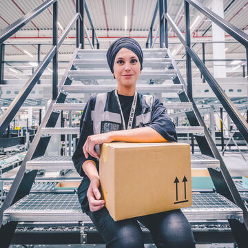 Arvato employee sits on a staircase in the warehouse, has her arm loosely resting on a package and smiles into the camera